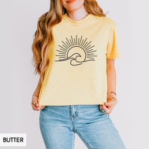 May include: A yellow cropped t-shirt with a black line drawing of a sun and waves. The shirt is worn by a person with long brown hair and blue jeans.