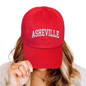 Puede incluir: Gorra de b&eacute;isbol roja con la palabra "ASHEVILLE" bordada en blanco en la parte delantera. La gorra tiene una visera curva y est&aacute; hecha de un tejido suave. Un sombrero de estilo cl&aacute;sico, adecuado para el uso diario.