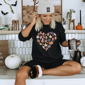 May include: A woman wearing a black sweatshirt with a heart-shaped design featuring autumn leaves, pumpkins, and acorns. She is holding a black mug with the text "Witchy Brew" and sitting on a counter with a white pumpkin and a sign that reads "Fall is my second favourite F-word".