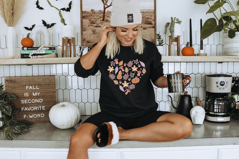 May include: A woman wearing a black sweatshirt with a heart-shaped design featuring autumn leaves, pumpkins, and acorns. She is holding a black mug with the text "Witchy Brew" and sitting on a counter with a white pumpkin and a sign that reads "Fall is my second favourite F-word".