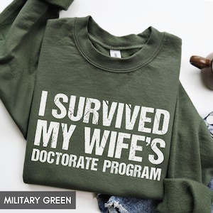 May include: Military green sweatshirt with the text "I SURVIVED MY WIFE'S DOCTORATE PROGRAM" in white block letters. The crew neck sweatshirt is made of a soft material and is folded on a white surface.