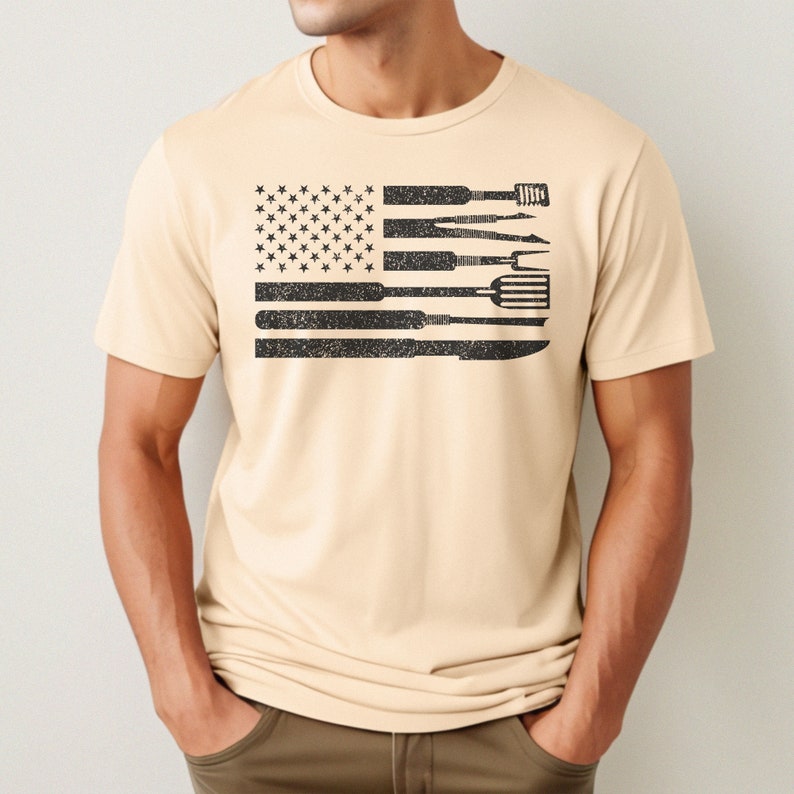 Puede incluir: Una camiseta beige con un dise&ntilde;o de bandera estadounidense en blanco y negro. La bandera est&aacute; compuesta por varias herramientas para asar a la parrilla, como una esp&aacute;tula, pinzas y un cepillo para asar.