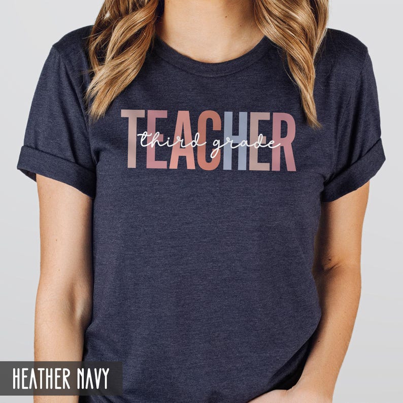 A woman modeling a heather navy t-shirt featuring multicolored block letters spelling teacher and white cursive text that says third grade.