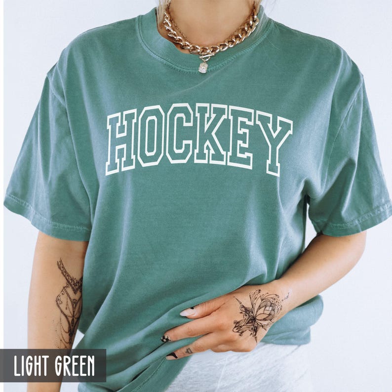 A woman wearing a light green t-shirt with white block letters spelling HOCKEY.