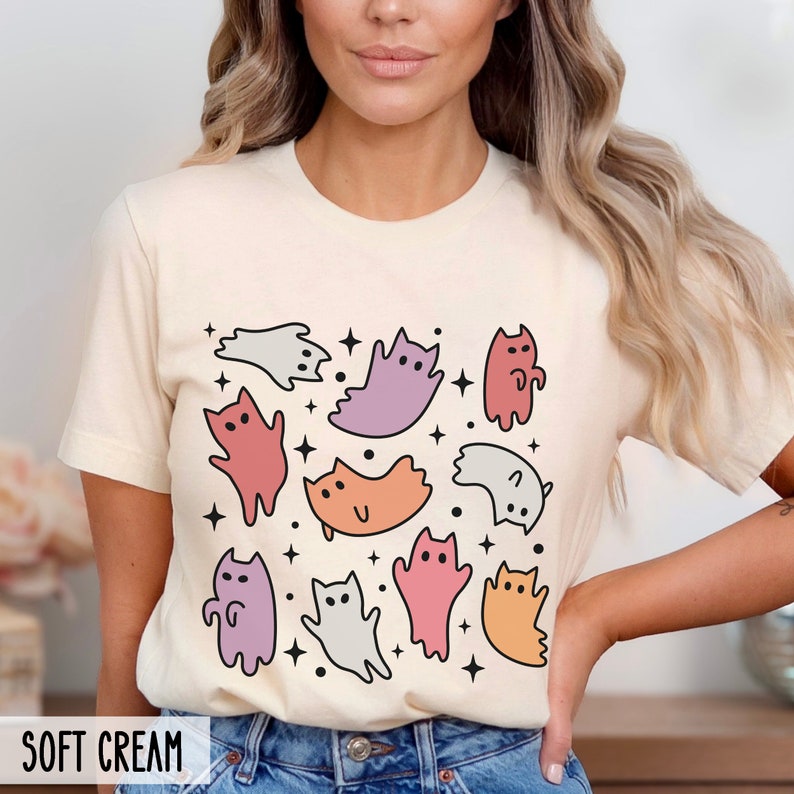 Op de afbeelding: Een cr&egrave;mekleurig T-shirt met een grafische print van kleurrijke cartoonkatten en sterren. De tekst "SOFT CREAM" is onderaan de afbeelding gedrukt.