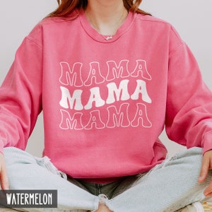 May include: A pink sweatshirt with the words "Mama Mama Mama" in white, retro-style lettering. The sweatshirt is made of a soft, comfortable material and has a relaxed fit.
