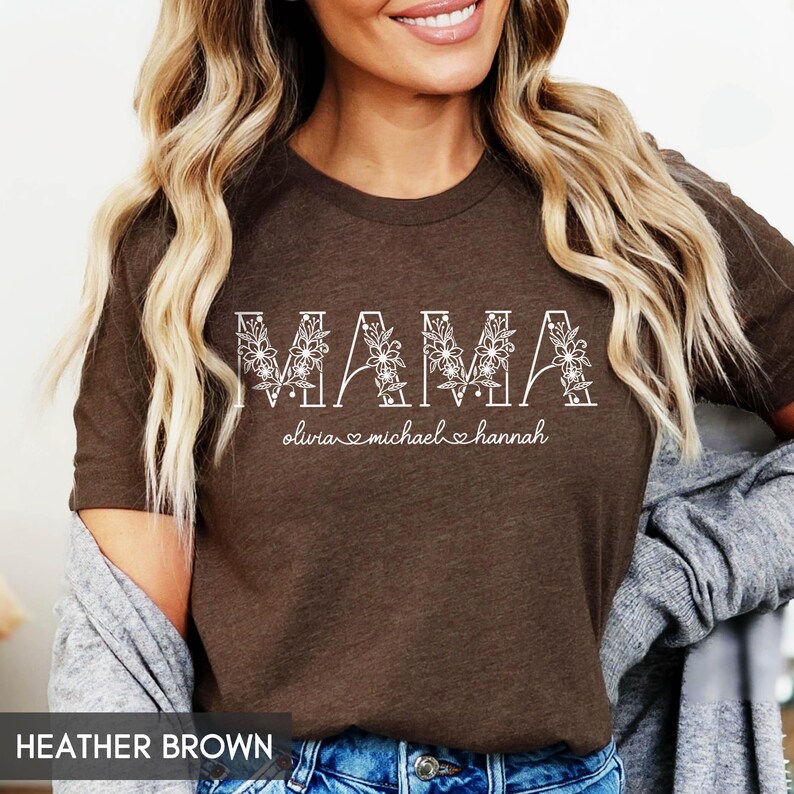 May include: Heather brown t-shirt with the word "MAMA" in white floral letters. Below the word "MAMA" are the names Olivia, Michael, and Hannah. The t-shirt is a crew neck style.