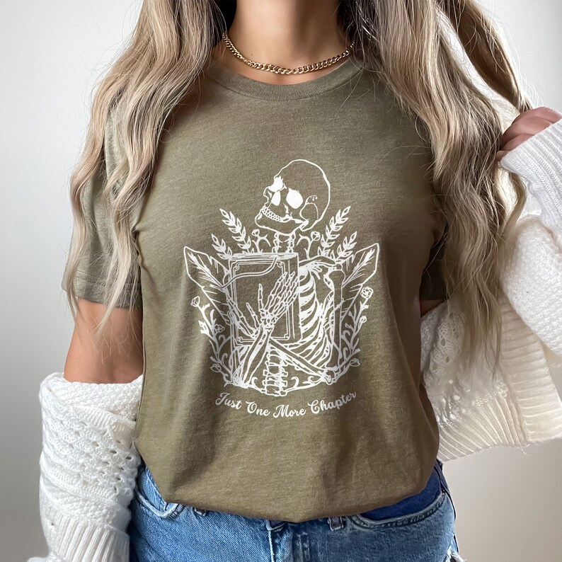 May include: A green t-shirt with a white graphic of a skeleton holding a book and surrounded by leaves. The text "Just One More Chapter" is printed below the graphic.