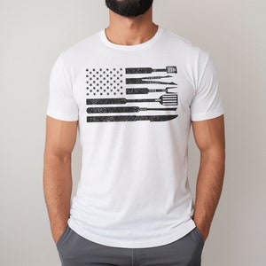 Puede incluir: Camiseta blanca con un dise&ntilde;o de bandera americana en blanco y negro. La bandera est&aacute; compuesta por herramientas, como una esp&aacute;tula, un cuchillo y un tenedor.