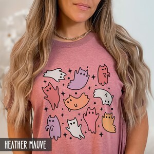 Op de afbeelding: Een heide-paarse t-shirt met een grafische print van kleurrijke katten en geesten. De grafiek bevat een verscheidenheid aan kleuren, waaronder roze, paars, oranje en wit.