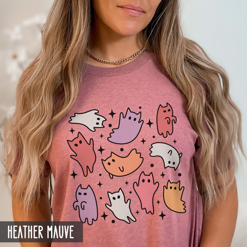 Op de afbeelding: Een heide-paarse t-shirt met een grafische print van kleurrijke katten en geesten. De grafiek bevat een verscheidenheid aan kleuren, waaronder roze, paars, oranje en wit.