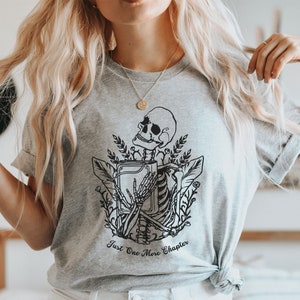 May include: A gray t-shirt with a black and white illustration of a skeleton holding a book. The text "Just One More Chapter" is printed below the illustration.