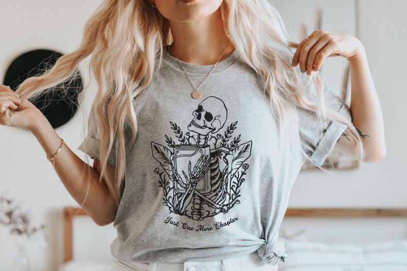 May include: A gray t-shirt with a black and white illustration of a skeleton holding a book. The text "Just One More Chapter" is printed below the illustration.