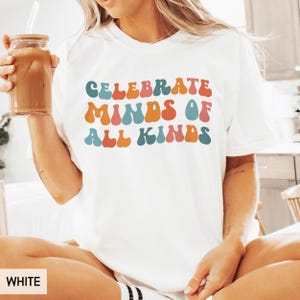May include: White t-shirt with the text "CELEBRATE MINDS OF ALL KINDS" in colorful bubble letters. The shirt is a casual style, perfect for everyday wear. The person in the image is holding a glass of iced coffee.