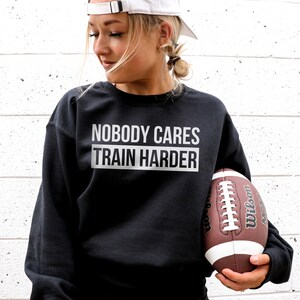 Pu&ograve; includere: Felpa nera con la scritta "Nobody Cares Train Harder" in lettere bianche. Una persona tiene un pallone da football marrone e bianco.