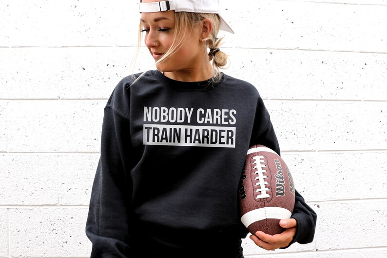 Pu&ograve; includere: Felpa nera con la scritta "Nobody Cares Train Harder" in lettere bianche. Una persona tiene un pallone da football marrone e bianco.