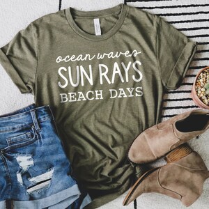 May include: A green t-shirt with white text that reads "ocean waves sun rays beach days". The shirt is laid out on a white and black striped surface with a pair of brown ankle boots and a potted cactus.