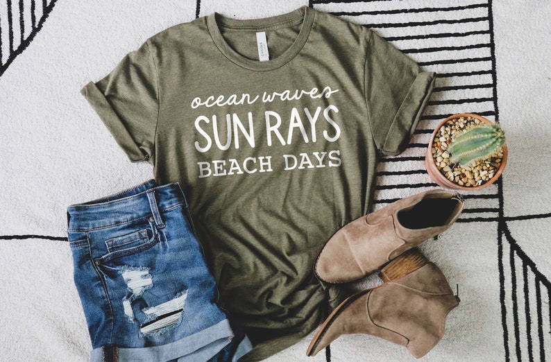 May include: A green t-shirt with white text that reads "ocean waves sun rays beach days". The shirt is laid out on a white and black striped surface with a pair of brown ankle boots and a potted cactus.