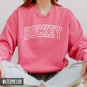 Puede incluir: Una sudadera rosa con la palabra "HOCKEY" impresa en blanco en la parte delantera. La sudadera est&aacute; hecha de un material suave y c&oacute;modo y tiene un corte holgado.