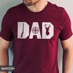 May include: Maroon t-shirt with the word "DAD" in white block letters. The letter "D" features a rifle, the "A" has a target, and the last "D" has a deer silhouette.