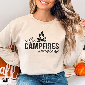A person wears a sand-colored crewneck sweatshirt featuring black text that reads coffee CAMPFIRES & cocktails with a campfire graphic.