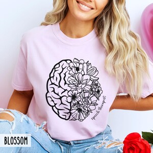May include: A pink t-shirt with a black and white design of a brain and flowers. The text "Speech Therapist" is written on the shirt.