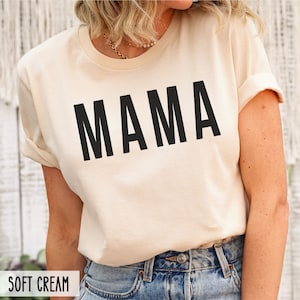 May include: A soft cream colored t-shirt with the word "MAMA" printed in black on the front.
