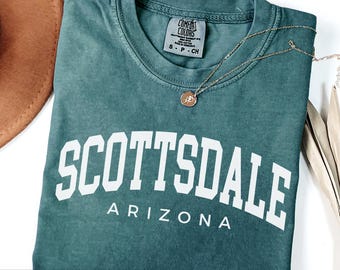 May include: A blue spruce t-shirt with the word "SCOTTSDALE" in large white letters, with "ARIZONA" below. The shirt is folded and has a small tag at the neckline. A gold necklace with a pendant is draped across the shirt.