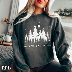 May include: A dark gray sweatshirt with a white graphic design of a forest silhouette with a full moon and the text "NORTH CAROLINA".
