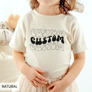 May include: A child wearing a natural colored t-shirt with the word "CUSTOM" repeated three times in a retro, black, bubble-letter font.