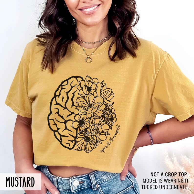 May include: Mustard yellow t-shirt with a black outline of a brain and flowers. The text "speech therapist" is written on the shirt.