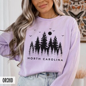 May include: A lavender long-sleeved sweatshirt with a black graphic of a forest silhouette and the text "NORTH CAROLINA".