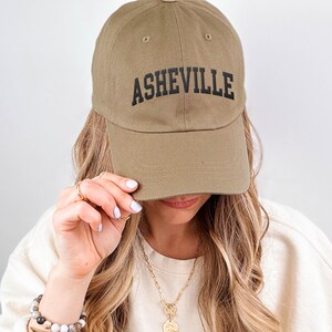 Puede incluir: Gorra de b&eacute;isbol de color caqui con la palabra "ASHEVILLE" impresa en negro en la parte delantera. La gorra est&aacute; hecha de una tela suave y tiene una visera curva. La gorra est&aacute; siendo sostenida por una persona.