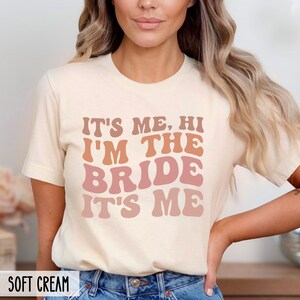 May include: A white t-shirt with the text "It's me, hi I'm the bride it's me" in a retro font with pink and orange color gradients.