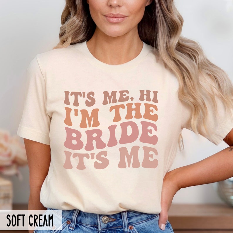 May include: A white t-shirt with the text "It's me, hi I'm the bride it's me" in a retro font with pink and orange color gradients.