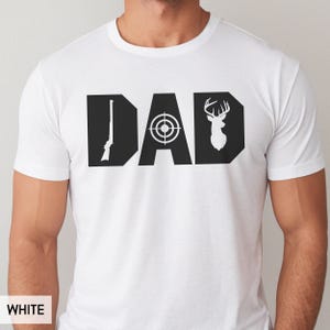 May include: White t-shirt with a black graphic design that spells out "DAD". The letter "D" features a rifle, the "A" has a target, and the final "D" has a deer silhouette. The shirt is made of a soft material.