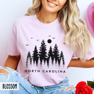 以下が含まれることがあります： 黒い森のシルエットと「NORTH CAROLINA」の文字が書かれたピンクのTシャツです。