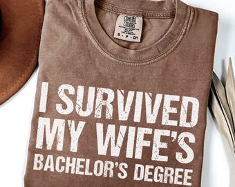 May include: Espresso-colored t-shirt with the text "I SURVIVED MY WIFE'S BACHELOR'S DEGREE" in white, distressed lettering. The shirt has a crew neck and a small tag. A brown hat is partially visible.