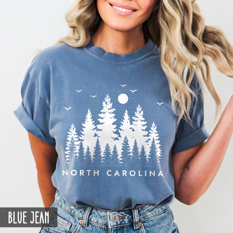 以下が含まれることがあります： 白い森のシルエットと「NORTH CAROLINA」の文字が書かれた、青いジーンズのTシャツです。