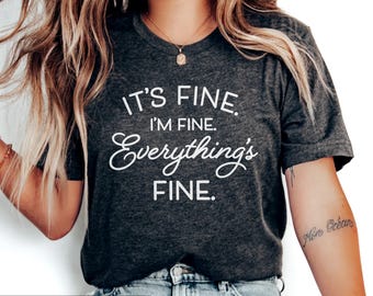 Puede incluir: Una camiseta de manga corta en color gris jaspeado oscuro con el texto en blanco que dice IT'S FINE. I'M FINE. Everything's FINE. La prenda tiene un cuello redondo y se combina con unos vaqueros de mezclilla azul claro.