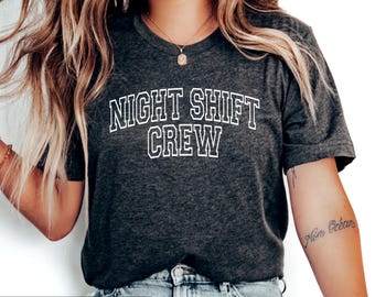 Może przedstawiać: Ciemnoszary melanżowy t-shirt z białym napisem NIGHT SHIFT CREW wykonanym czcionką w stylu uniwersyteckim. Koszulka ma krótkie rękawy i okrągły dekolt. Napis znajduje się na środku klatki piersiowej i jest zestawiony z niebieskim dżinsem.
