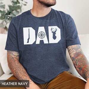 May include: Heather navy t-shirt with the word "DAD" in white block letters. The "D" features a rifle, the "A" has a target, and the second "D" has a deer silhouette. The shirt is made of a soft material.