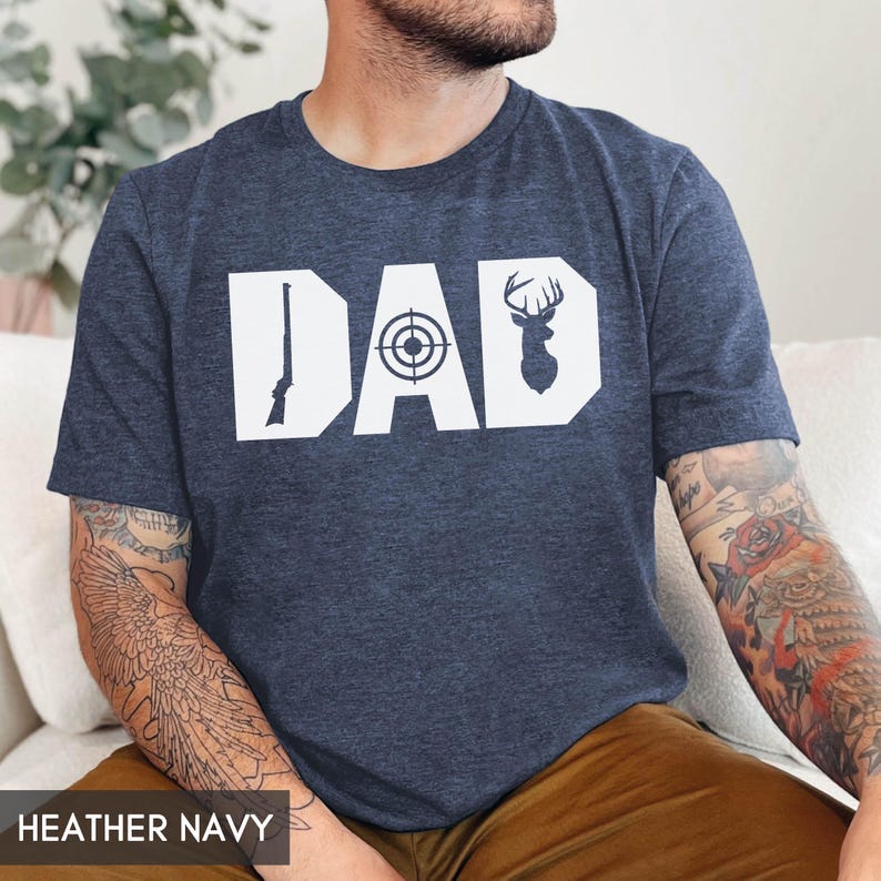 May include: Heather navy t-shirt with the word "DAD" in white block letters. The "D" features a rifle, the "A" has a target, and the second "D" has a deer silhouette. The shirt is made of a soft material.