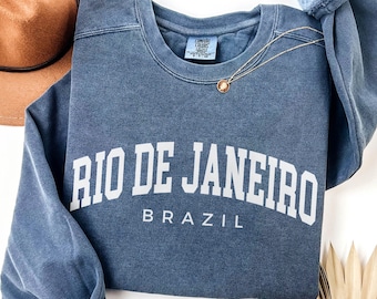 Peut inclure: Un sweat-shirt bleu denim avec l'inscription "RIO DE JANEIRO" en lettres blanches, avec "BRAZIL" en dessous. Un collier doré avec un pendentif repose sur le sweat-shirt. Un chapeau marron est visible en arrière-plan.