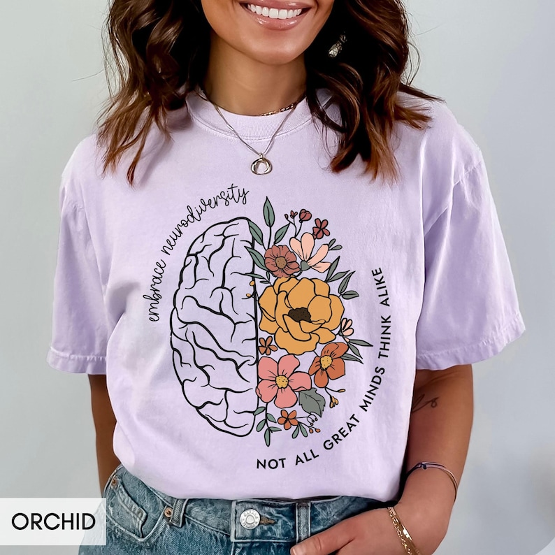 以下が含まれることがあります： 片側に花がついた脳のグラフィックと、反対側に「Embrace Neurodiversity Not All Great Minds Think Alike」というテキストが書かれたパープルのTシャツです。