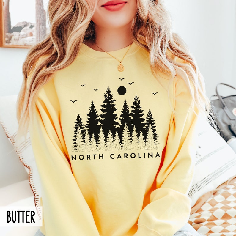 May include: A yellow sweatshirt with a black graphic of a forest silhouette, a moon, and birds. The text "NORTH CAROLINA" is printed below the graphic.