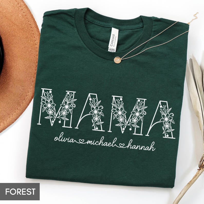May include: Forest green t-shirt with the word "MAMA" in white floral lettering. Below the word "MAMA" are the names "olivia", "michael", and "hannah" with heart symbols. A gold necklace and a brown hat are also visible.
