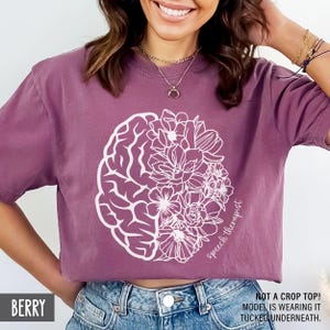 May include: A purple t-shirt with a white outline of a brain and flowers. The text "Speech therapist" is written on the shirt.