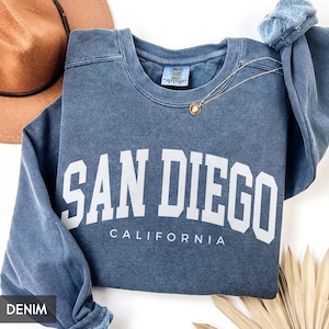 May include: A denim blue sweatshirt with the words "SAN DIEGO CALIFORNIA" in white block letters. A gold necklace with a pendant is draped across the sweatshirt. A brown hat is visible in the background.