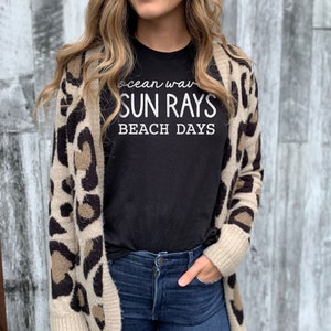 May include: A woman wearing a black t-shirt with the text "ocean waves sun rays beach days" printed on it. She is wearing a brown and white leopard print cardigan and blue jeans.